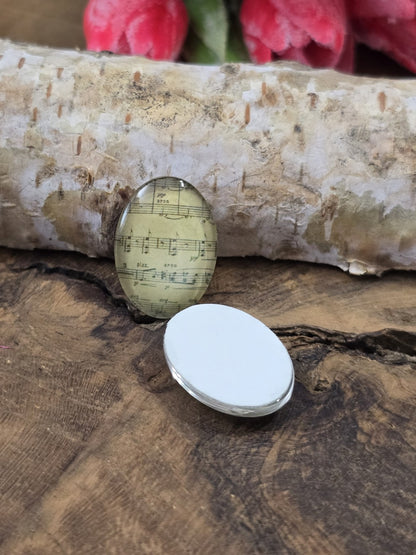 Zwei ovale Glascabochons mit Musiknoten auf Holz, wovon einer mit der flachen, weißen Rückseite nach oben liegt, um die Klebefläche für Fassungen zu zeigen.