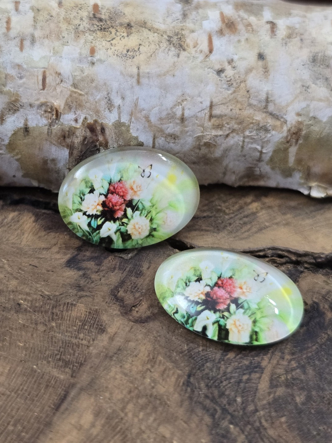 Zwei ovale Glascabochons (18x13 mm) mit nostalgischem Blumenstrauß-Motiv und kleinem Schmetterling auf rustikalem Holz vor einem hellen Birkenstamm.