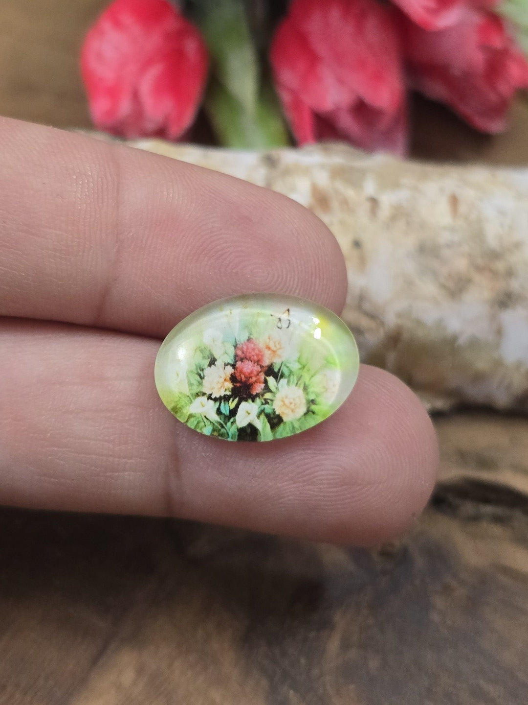 Ein ovaler Vintage-Glascabochon (18x13 mm) mit buntem Blumenmotiv auf einer Fingerspitze als Größenvergleich für DIY-Schmuckprojekte.