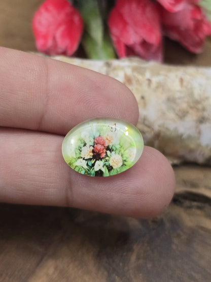 Ein ovaler Vintage-Glascabochon (18x13 mm) mit buntem Blumenmotiv auf einer Fingerspitze als Größenvergleich für DIY-Schmuckprojekte.