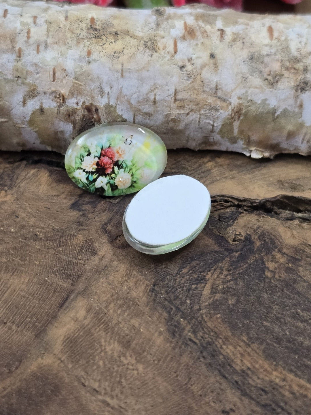 Zwei ovale Blumen-Glascabochons auf Holz, wovon einer mit der flachen, weißen Rückseite nach oben liegt, um die Klebefläche für Fassungen zu zeigen.