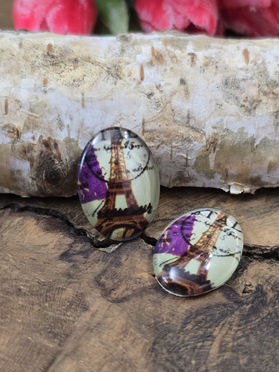Zwei ovale Glascabochons (18x13 mm) mit Eiffelturm-Motiv, Poststempel und lila Akzenten dekorativ platziert auf rustikalem Holz vor einem hellen Birkenstamm.