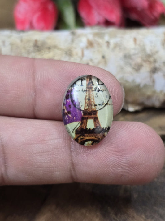 Ein ovaler Vintage-Glascabochon (18x13 mm) mit Eiffelturm und Motiv-Details in Paris-Optik auf einer Fingerspitze als praktischer Größenvergleich für DIY-Schmuck.