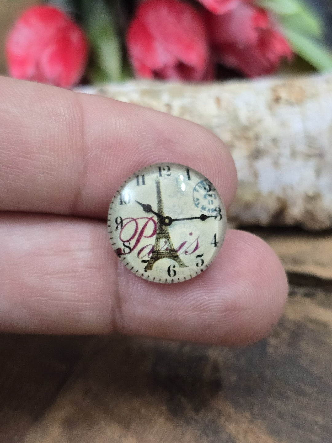 Ein runder Eiffelturm-Uhr-Glascabochon (16 mm) im nostalgischen Vintage-Stil auf einer Fingerspitze präsentiert als praktischer Größenvergleich.