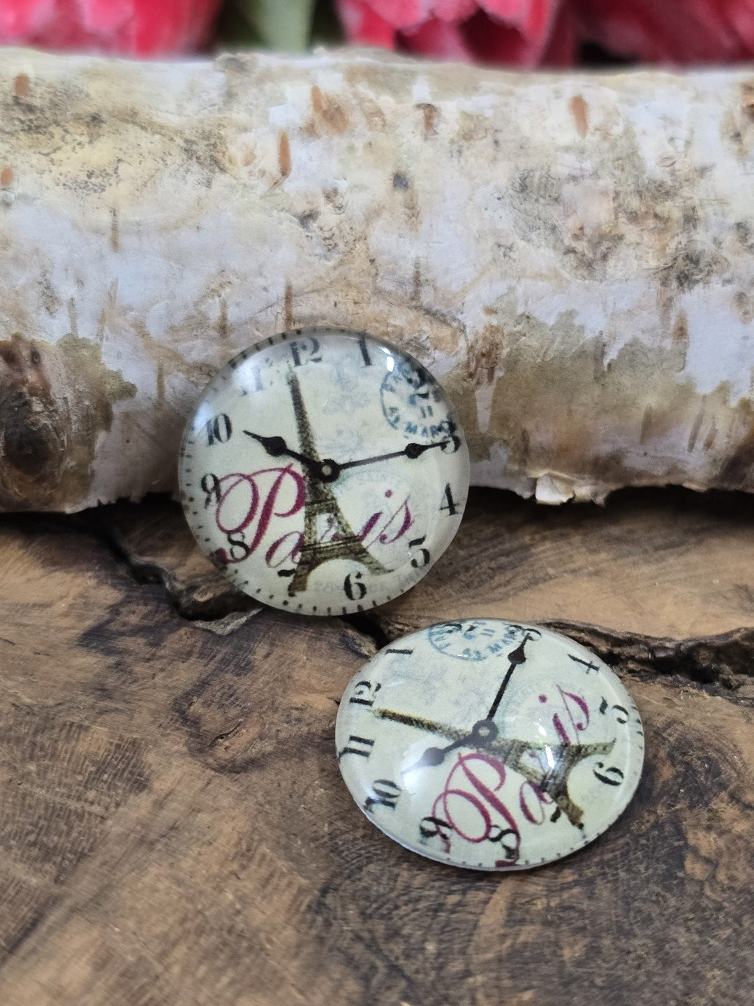 Zwei Paris-Schmucksteine aus Glas mit filigranem Uhr- und Eiffelturm-Motiv dekorativ arrangiert auf Holz vor einem hellen Birkenast.