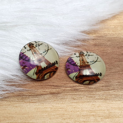 Zwei runde Glascabochons (16 mm) mit Eiffelturm-Motiv, Poststempel und lila Blüten im Vintage-Look auf rustikalem Holz vor weißem Kunstfell.