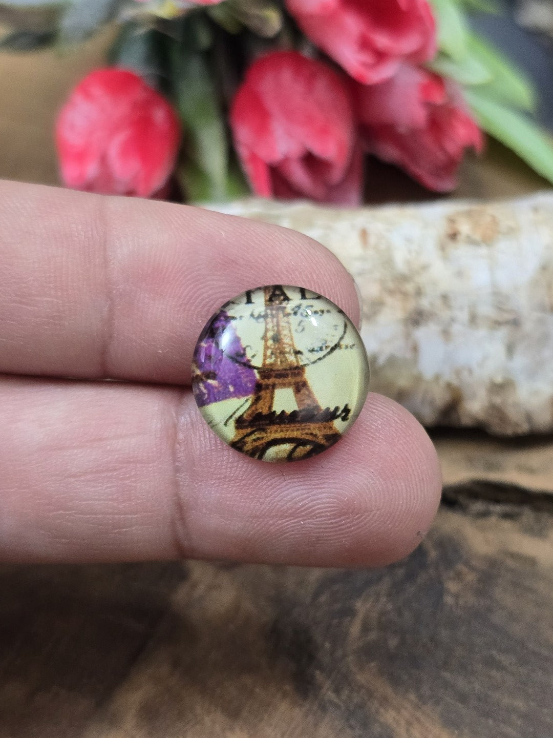Ein kleiner Vintage-Glascabochon (16 mm) mit Eiffelturm- und Blumen-Motiv auf einer Fingerspitze als praktischer Größenvergleich für DIY-Schmuckprojekte.