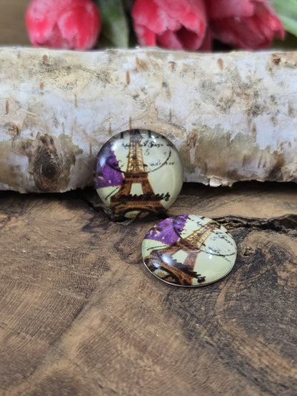 Zwei Eiffelturm-Schmucksteine aus Glas mit violetten Akzenten dekorativ arrangiert auf einer dunklen Holzoberfläche vor einem hellen Birkenstamm.