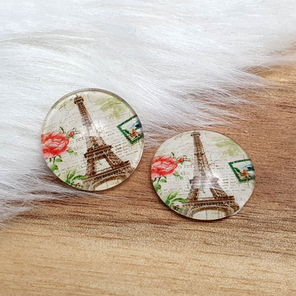 Zwei runde Glascabochons (16 mm) mit Vintage-Paris-Motiv, Eiffelturm, einer roten Rose und Poststempel auf rustikalem Holz vor weißem Fell.