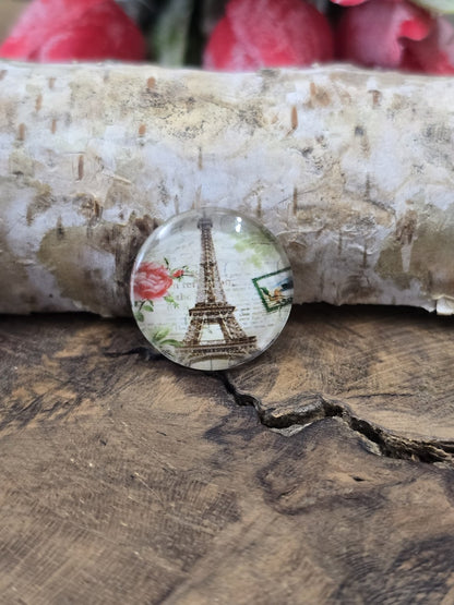 Nahaufnahme eines einzelnen runden Cabochons aus Glas mit Eiffelturm-Motiv vor Birkenrinde als Zubehör für die eigene Schmuckherstellung.