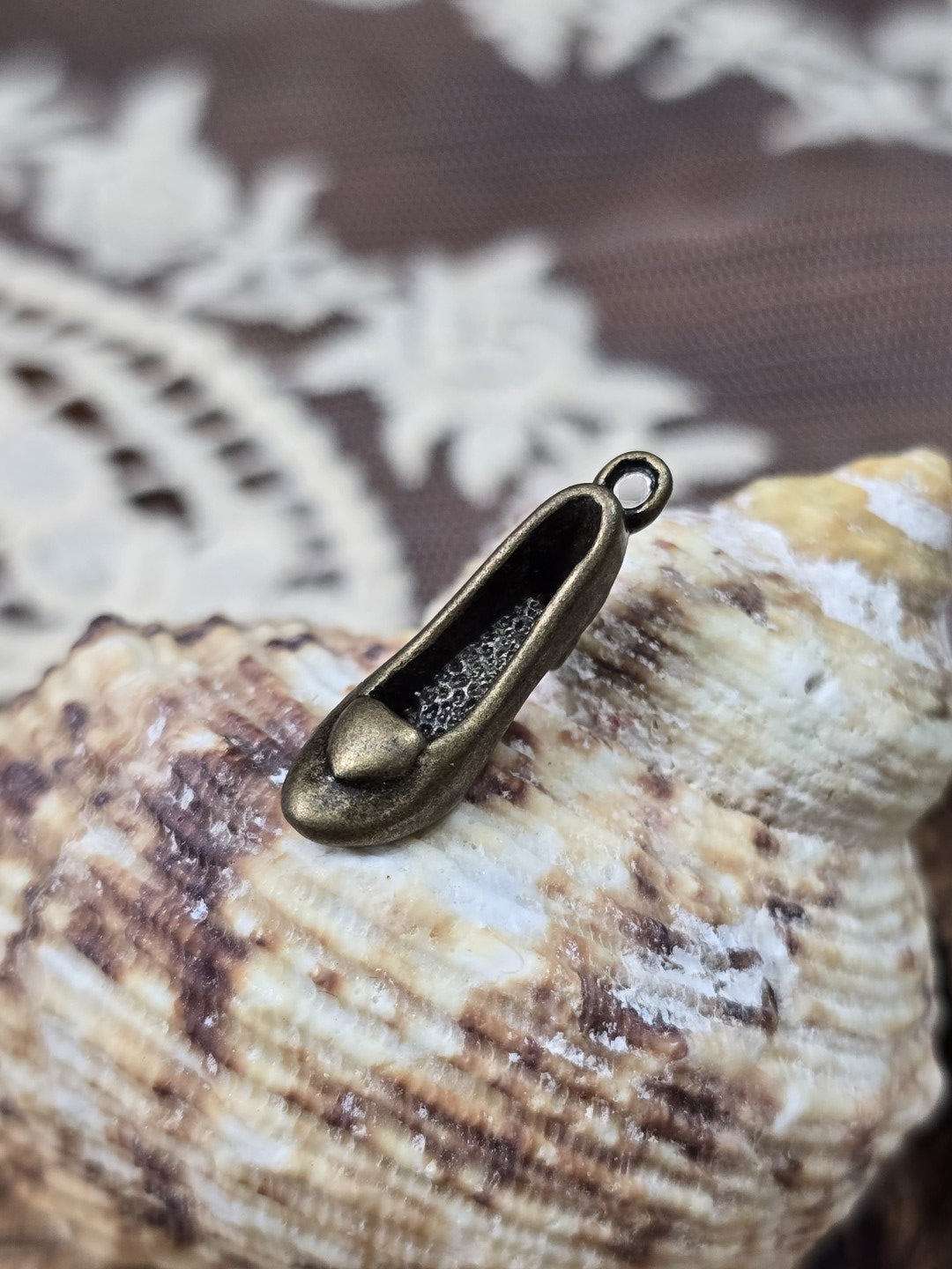 Schuh-Schmuckanhänger in Bronze-Optik mit Öse schräg vor einer Muschel dekoriert.