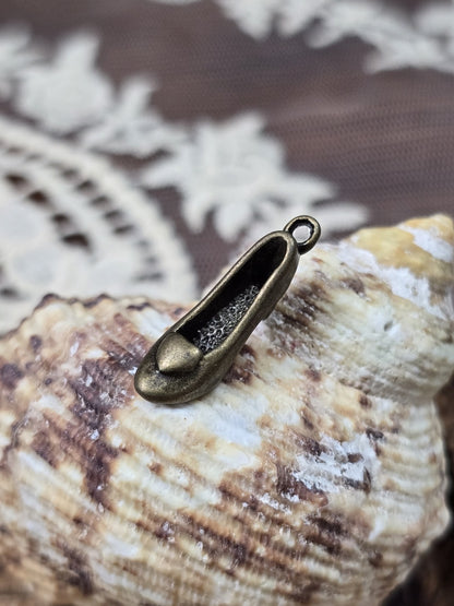 Schuh-Schmuckanhänger in Bronze-Optik mit Öse schräg vor einer Muschel dekoriert.