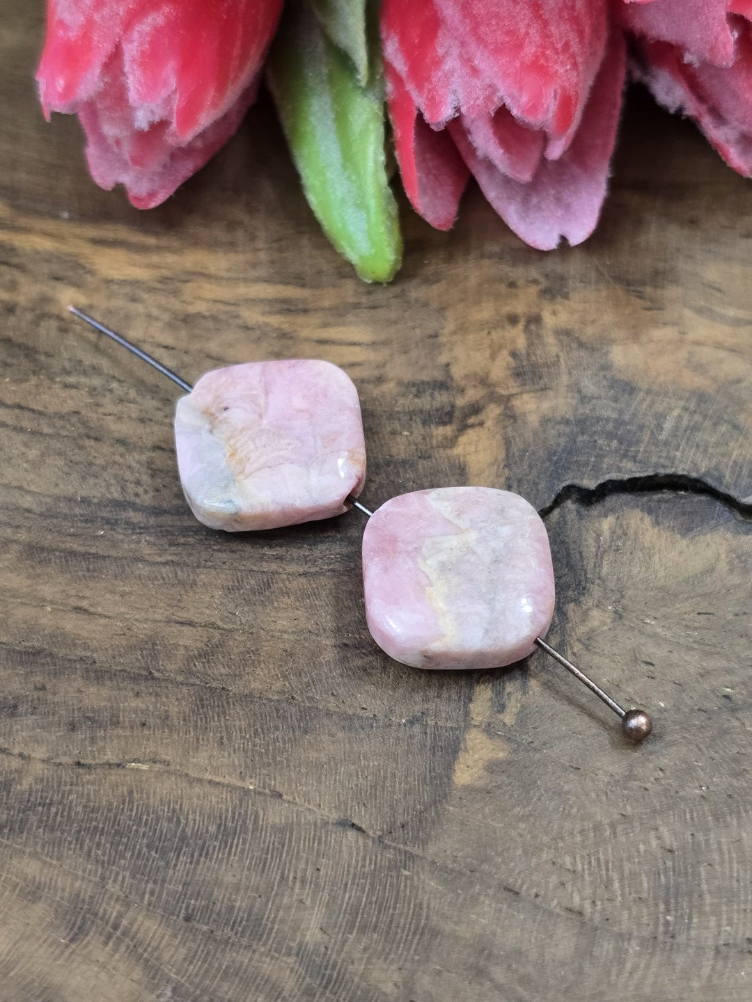 Zwei rosa Rhodochrosit-Edelsteinperlen (13,6 mm) in flacher Diamantform auf einen Metallstift aufgereiht vor dekorativen Blüten.