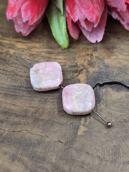 Zwei rosa Rhodochrosit-Edelsteinperlen (13,6 mm) in flacher Diamantform auf einen Metallstift aufgereiht vor dekorativen Blüten.