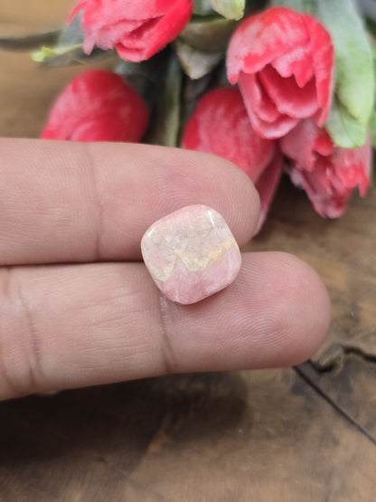 Eine quadratisch-abgerundete Rhodochrosit-Perle (13,6 mm) in Rosa-Beige auf der Handfläche als Größenvergleich.