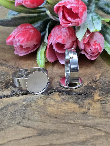 Silberfarbene verstellbare Ringschienen mit runder Fassung für 12mm Cabochons und gezacktem Rand vor rosa Blütendekoration.