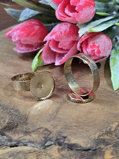 Goldene verstellbare Ringrohlinge mit runder 12mm Cabochon-Fassung für DIY-Schmuck vor rosa Blumen arrangiert.
