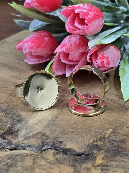 Goldene verstellbare Ringrohlinge mit runder 18mm Cabochon-Fassung und gezacktem Rand vor rosa Deko-Blumen.