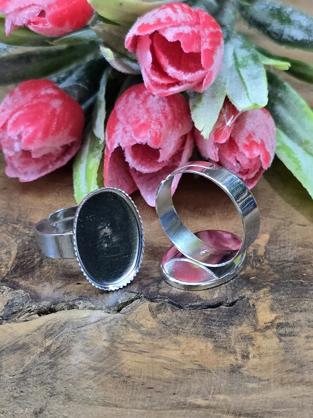 Silberfarbene verstellbare Ringschienen mit ovaler Klebefläche für Cabochons stilvoll vor rosa Blütendekoration arrangiert.