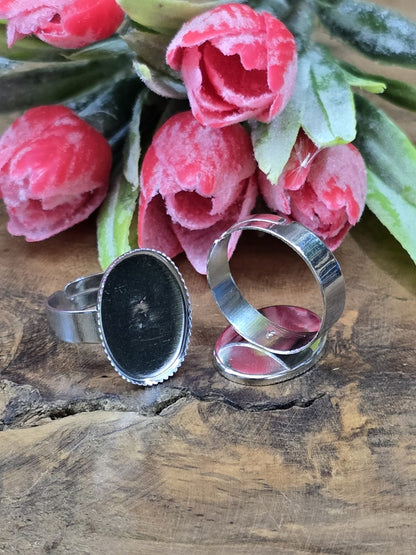 Silberfarbene verstellbare Ringschienen mit ovaler Klebefläche für Cabochons stilvoll vor rosa Blütendekoration arrangiert.