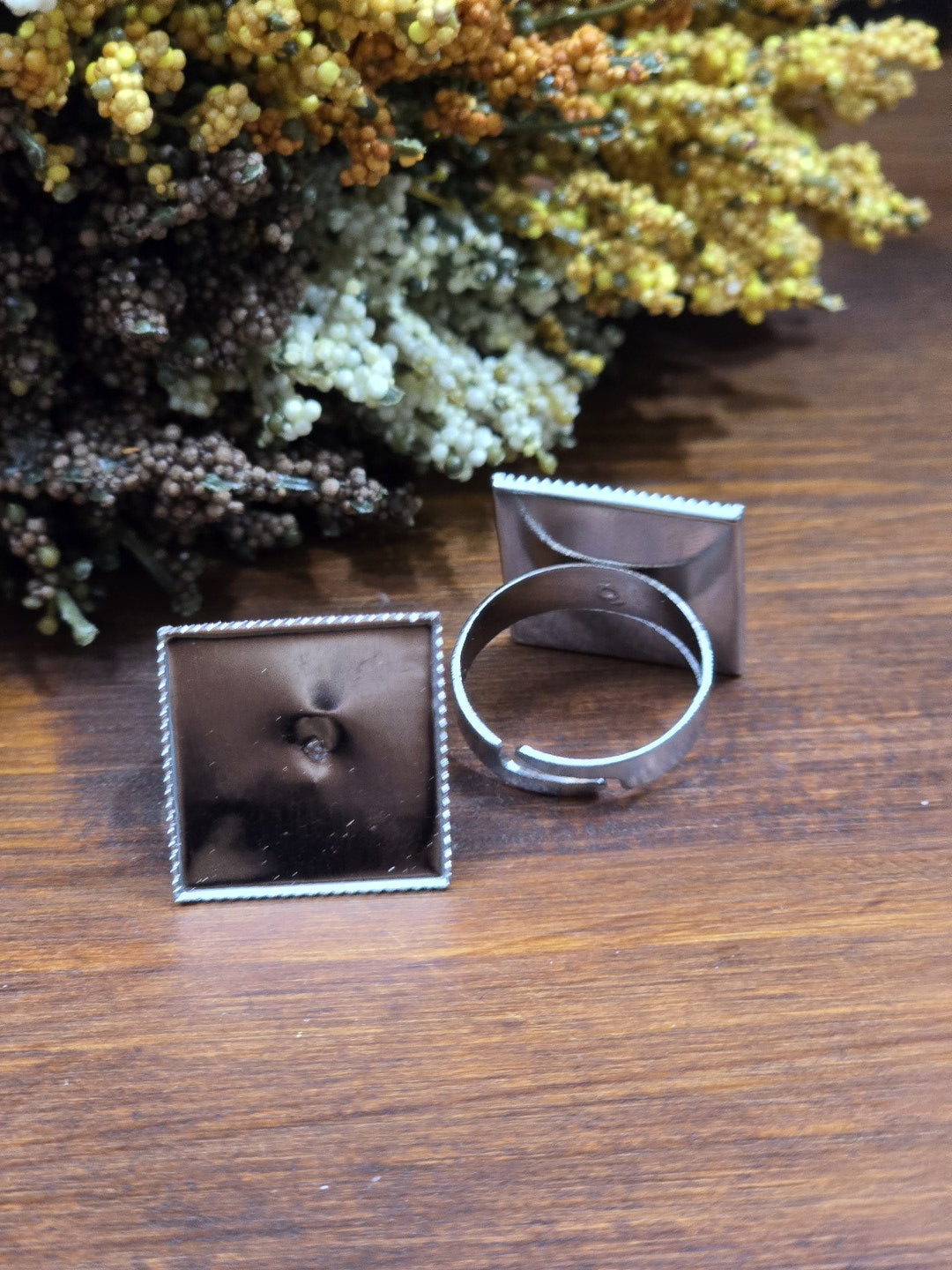 Verstellbare silberfarbene Ringschienen mit quadratischer 19-mm-Klebefläche und strukturiertem Rand vor getrockneten Blüten.