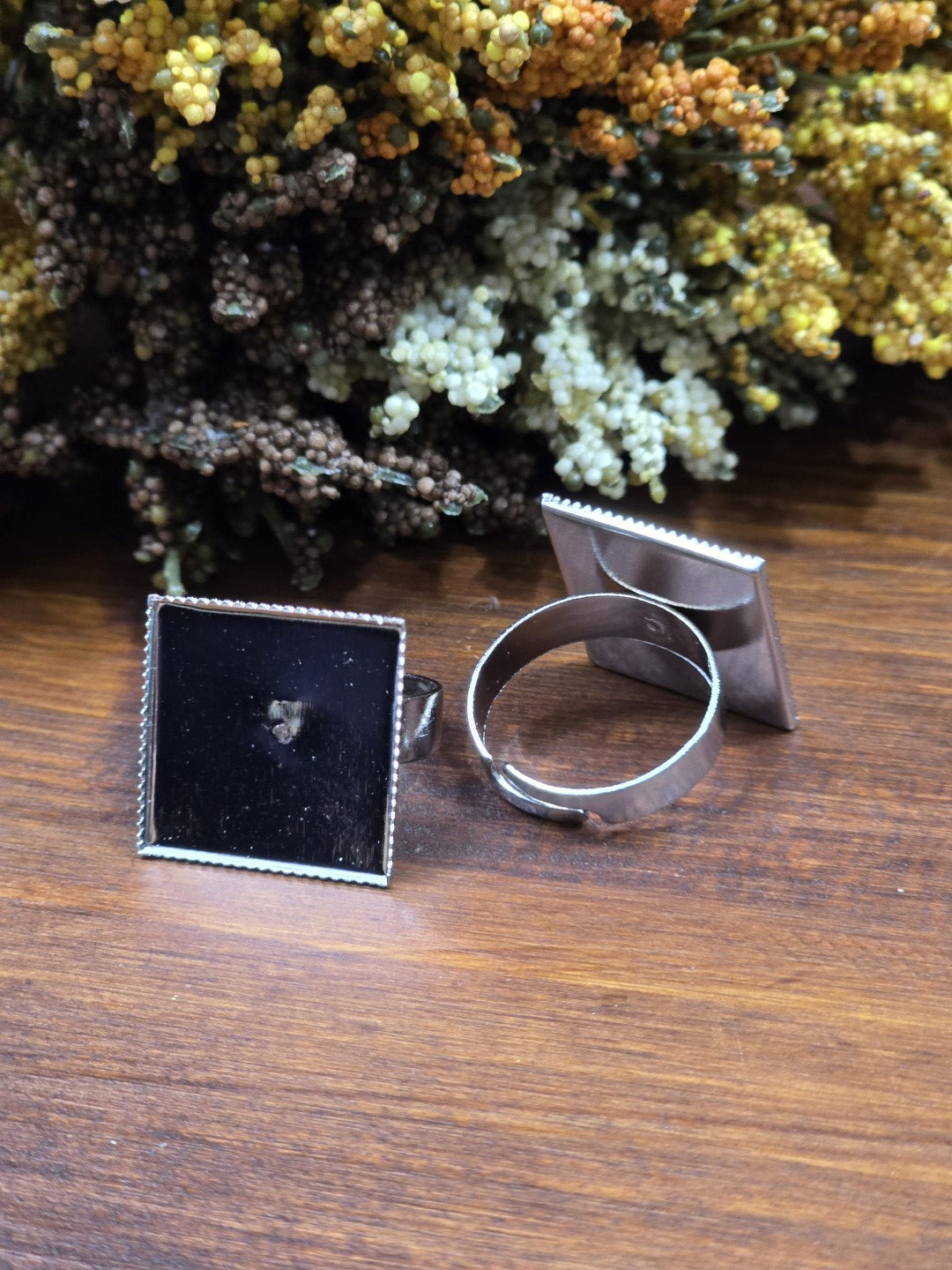 Zwei silberne verstellbare Ringrohlinge mit quadratischer Platte (19 mm) zur Schmuckherstellung vor floralem Hintergrund.
