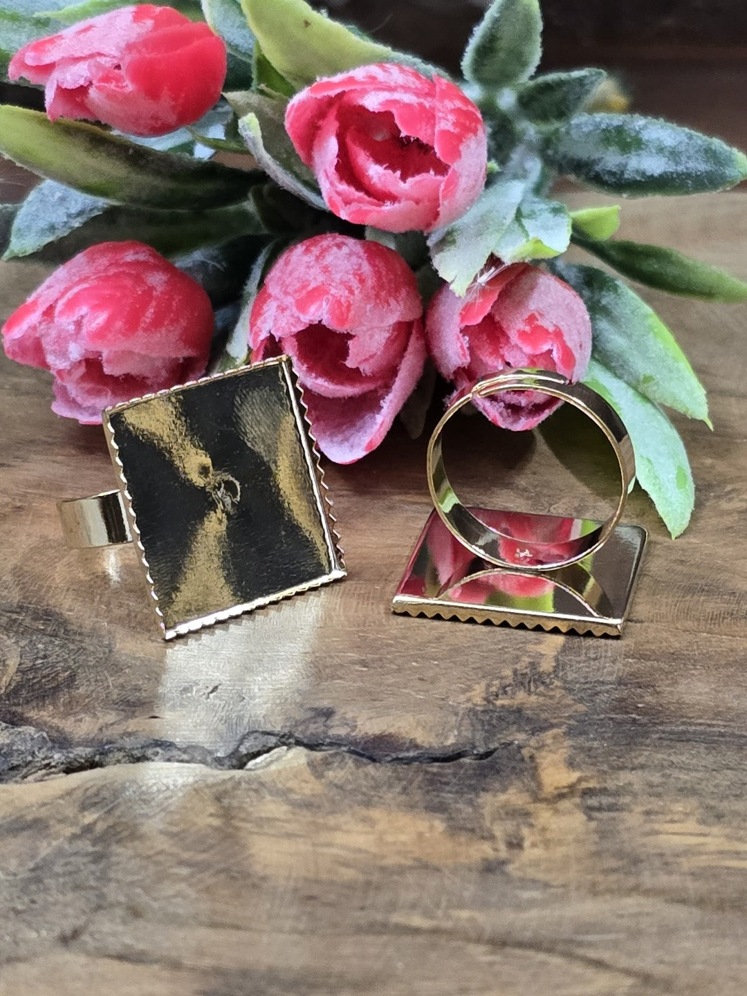 Zwei goldene Ringrohlinge mit quadratischer Klebefläche (19 mm) zur Schmuckherstellung auf Holz mit Blüten-Dekoration.