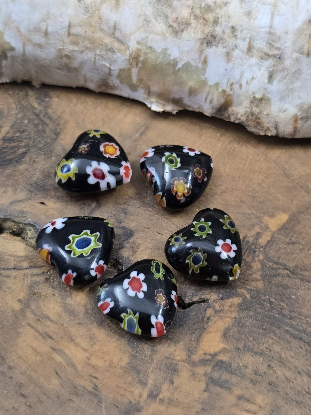 Fünf herzförmige schwarze Millefiori-Glasperlen mit buntem Blumenmuster kreisförmig auf Holzgrund platziert.
