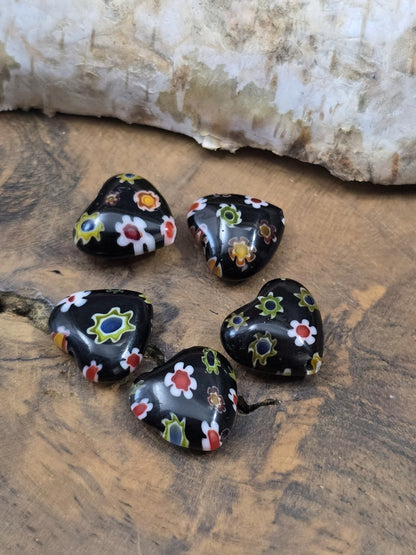 Fünf herzförmige schwarze Millefiori-Glasperlen mit buntem Blumenmuster kreisförmig auf Holzgrund platziert.