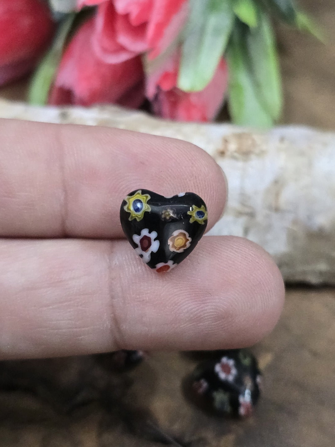 Eine schwarze Millefiori-Glasperle in Herzform mit Blütenmuster auf der Fingerkuppe als Größenvergleich.