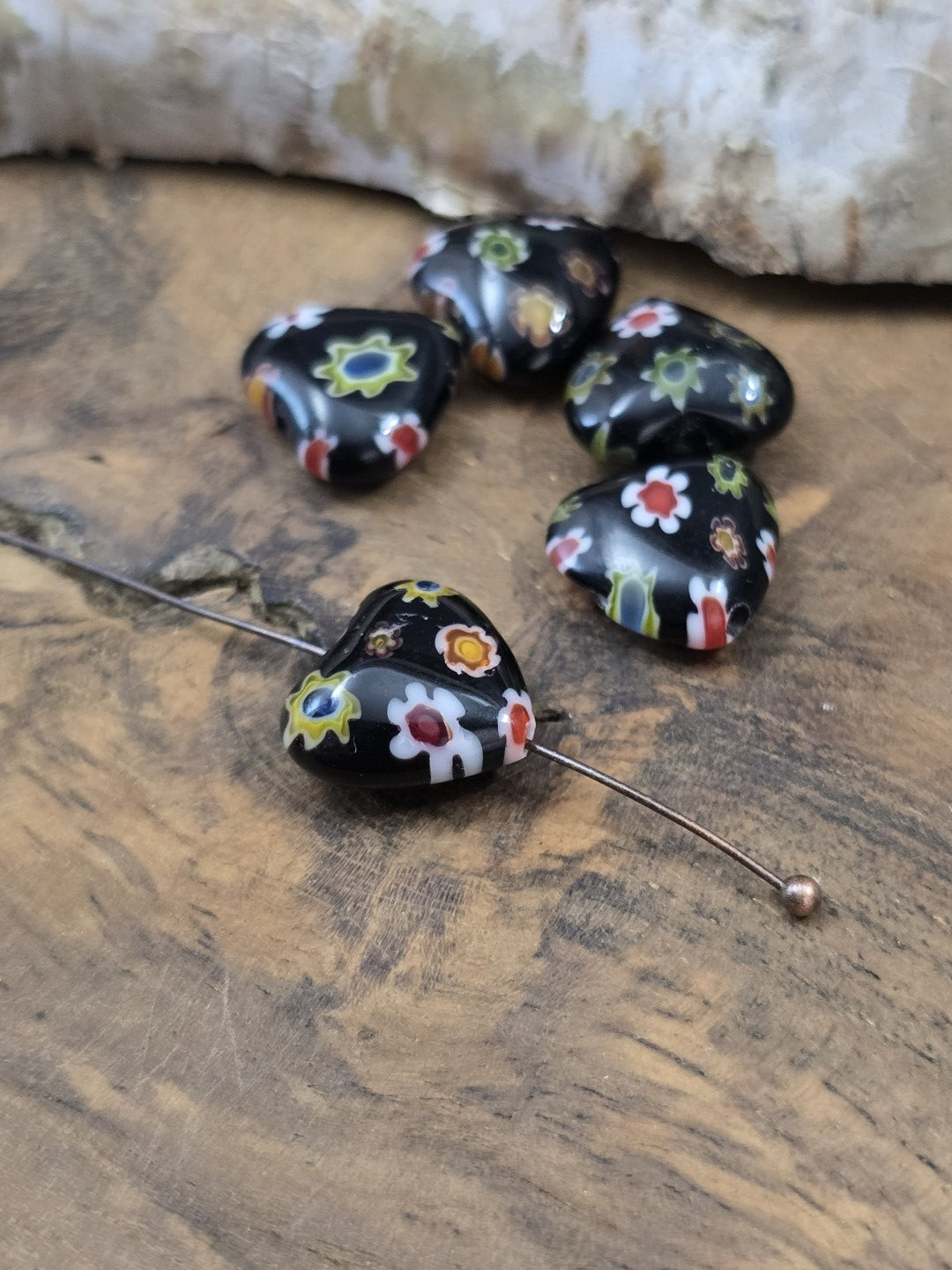 Schwarze Millefiori-Herzperle aus Glas auf einen Kugelnietstift aufgereiht neben vier weiteren Perlen zur Veranschaulichung der Bohrung.