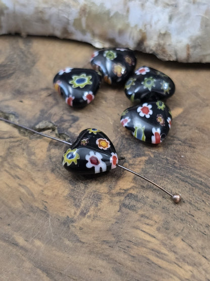 Schwarze Millefiori-Herzperle aus Glas auf einen Kugelnietstift aufgereiht neben vier weiteren Perlen zur Veranschaulichung der Bohrung.