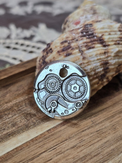 Runder Antik-Silber Anhänger im Uhrwerk-Design platziert vor einer Muschel auf Holz.