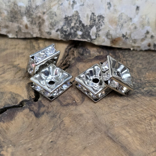 Vier eckige, silberfarbene Perlen mit funkelnden, klaren Strasssteinen aus Glas. Jede Seite der 8 x 8 mm großen Perle ist mit zwei Strasssteinen verziert. Perfekt für Armbänder, Ketten oder elegante Schmuckakzente. Fädelloch: 1,5 mm.