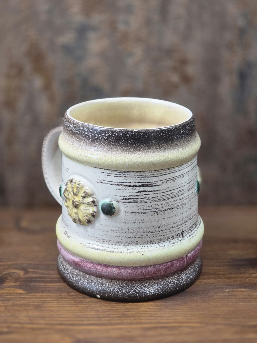 Vintage Keramikbecher mit hellem Korpus, seitlichem Blütenrelief und rosa-gelbem Sockelband in schräger Rückansicht