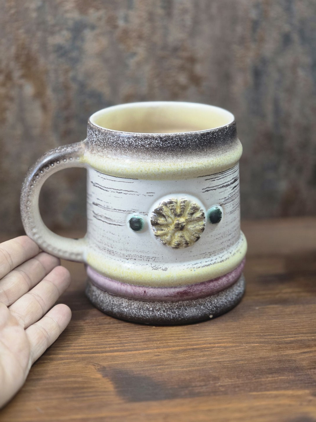 Vintage Keramikbecher mit hellem Korpus, gelbem Blütenmotiv und dunklem Rand neben einer Hand als Größenvergleich