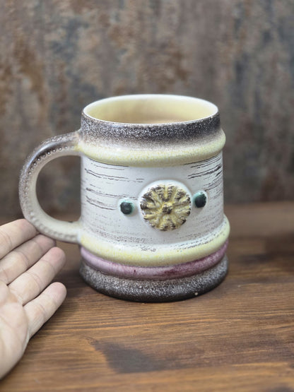 Vintage Keramikbecher mit hellem Korpus, gelbem Blütenmotiv und dunklem Rand neben einer Hand als Größenvergleich