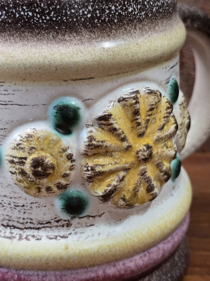 Vintage Keramikbecher mit floralem Reliefdekor in Gelb und Braun sowie grünen Punkten auf heller Oberfläche in Nahaufnahme