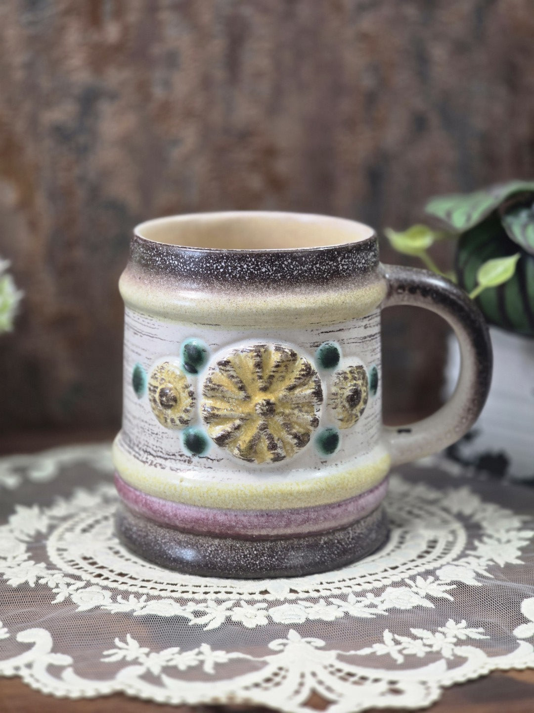 Vintage Keramikbecher mit gelbem Blütenrelief, grünen Akzenten und mehrfarbigen Glasurbändern auf dekorativer Spitzendecke