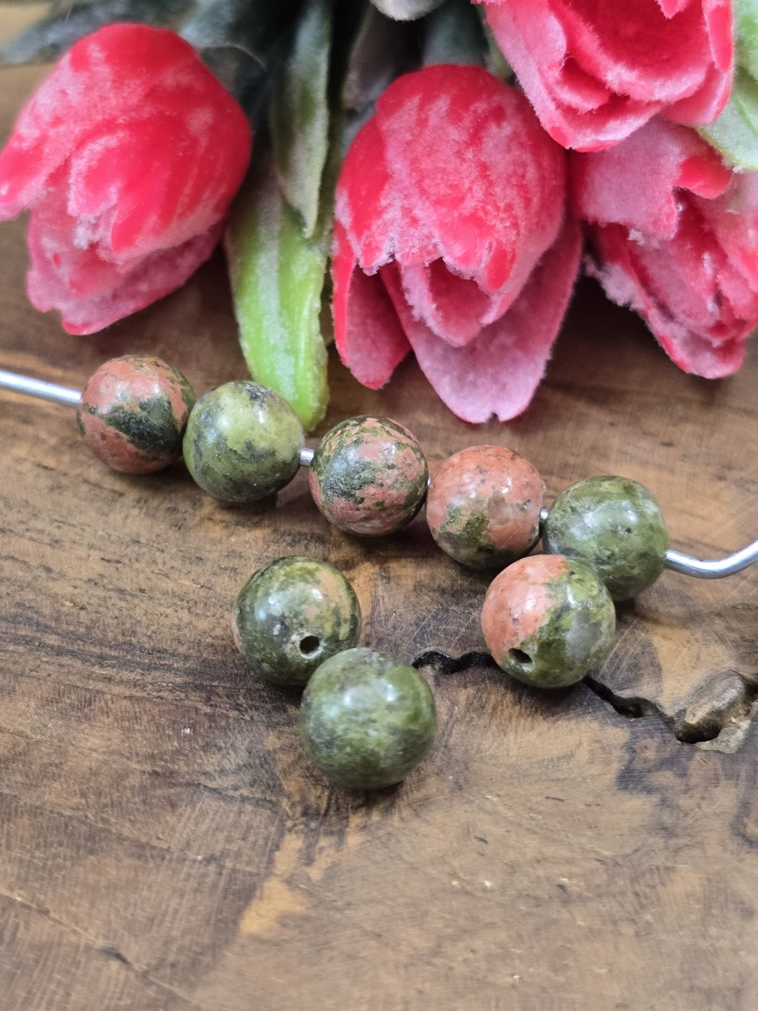 Acht Unakit-Edelsteinperlen (8 mm) in charakteristischem Rosa-Grün auf Draht und Holzgrund vor roten Blüten.