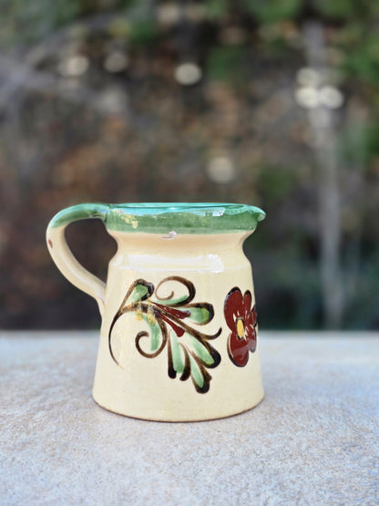 Vintage Keramikkanne mit cremefarbener Oberfläche, grünem Rand, handgemalter Blume und unglasiertem rotscherbigem Standboden.