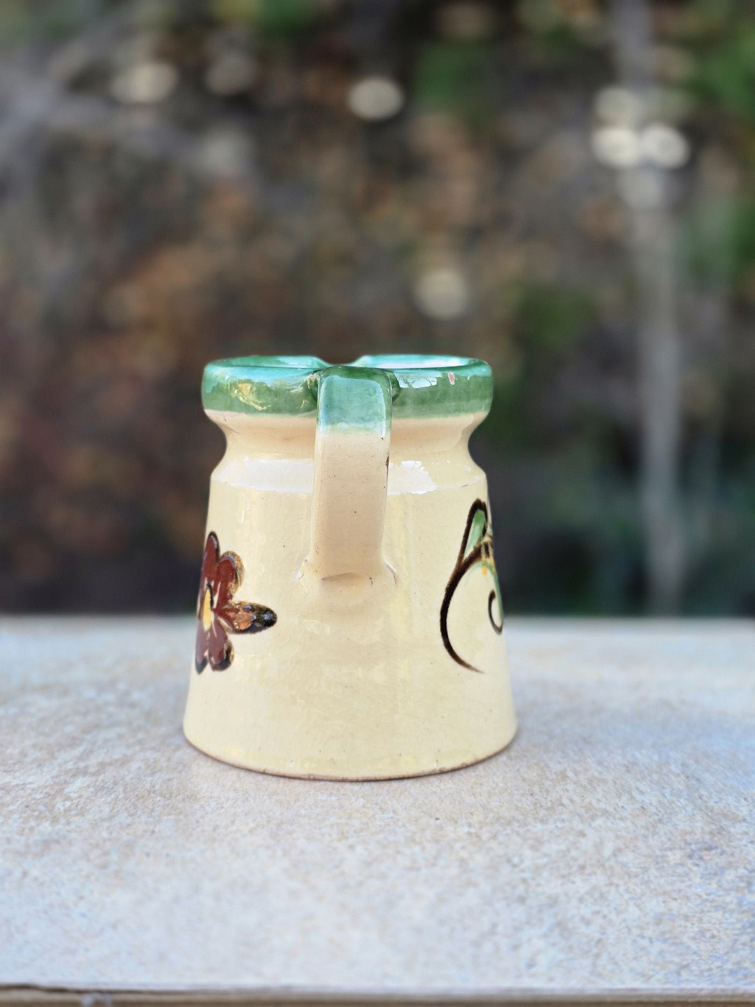 Kleine Majolika-Kanne im Vintage-Stil mit hellem Cremegrund, grünem Dekorrand, floraler Handbemalung und rotem Keramikscherben am Boden.