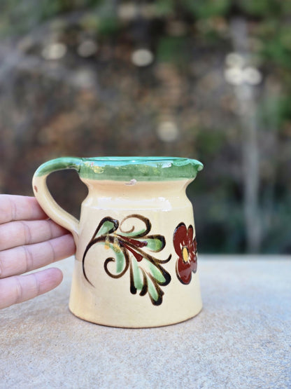 Vintage-Kanne aus glasierter Keramik mit Majolika-Charakter, cremefarbenem Grundton, grünem Rand, handgemalter Blume und rotscherbigem Boden.