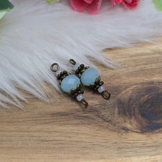 Das Bild zeigt zwei Verbinder mit mintfarbenen Kristallglasperlen und antik bronzefarbigen Metallelementen. Die Verbinder haben eine Länge von 22 mm und präsentieren sich mit einem eleganten, facettierten Design, das das Licht auf funkelnde Weise reflektiert. Die mintfarbenen Glasperlen strahlen eine subtile Frische aus, während die bronzefarbigen Elemente einen warmen, nostalgischen Akzent setzen. Die Kombination aus sanften Farben und hochwertigen Materialien macht diese Verbinder zu einem perfekte