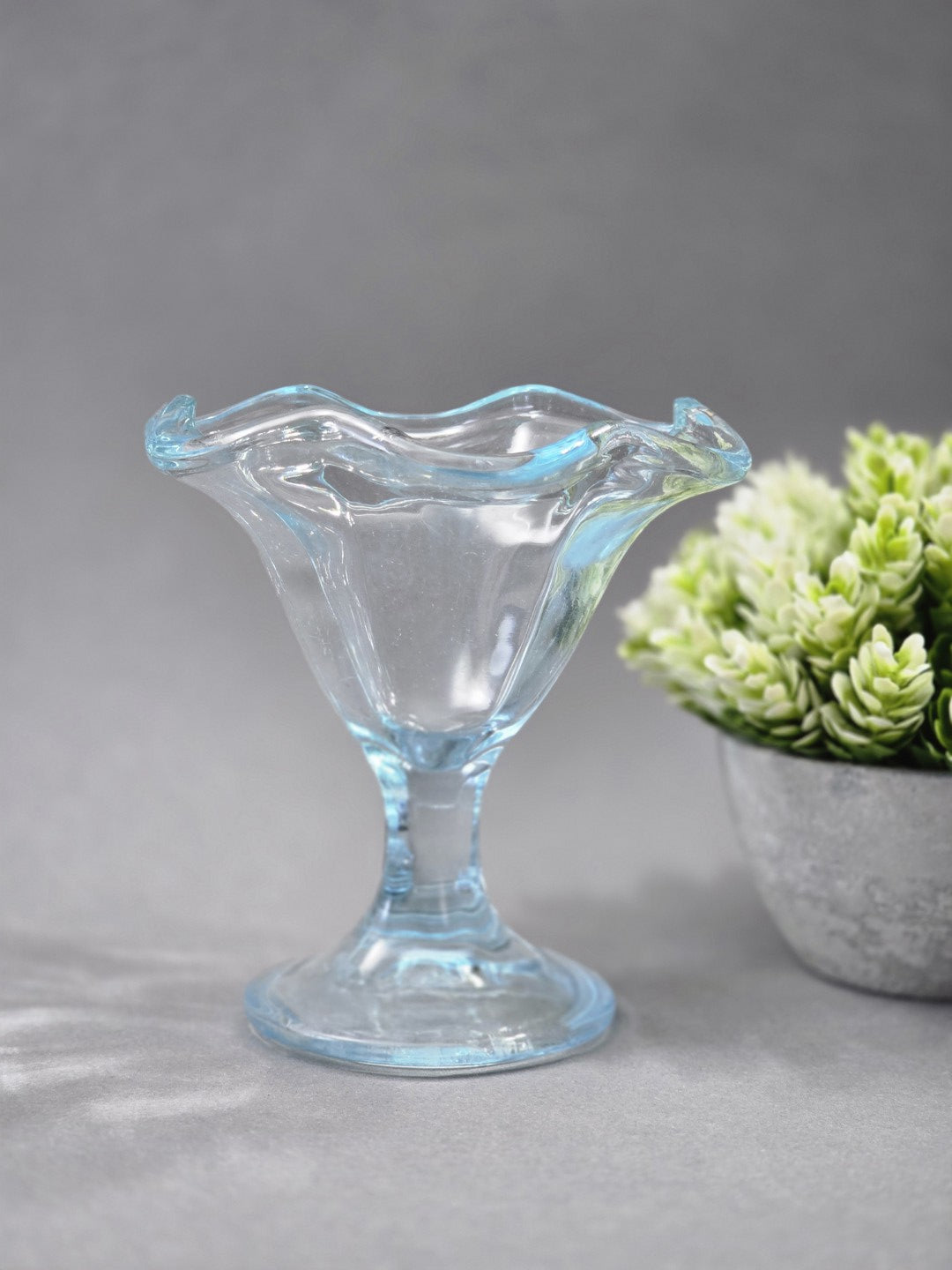 Ein transparenter Eisbecher aus hellblau getöntem Glas steht auf grauem Untergrund, mit gewelltem Rand, schmalem Stiel und rundem Fuß.