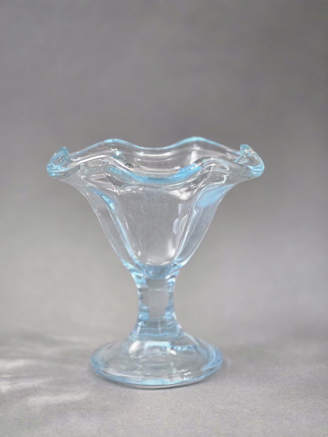 Der Eisbecher ist frontal zu sehen; die klare Schale öffnet sich nach oben wie eine Blüte und der Rand schimmert zart hellblau.