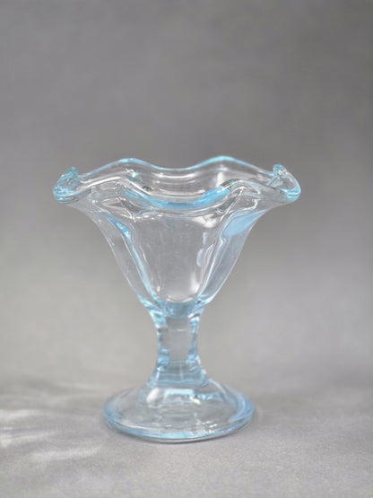 Der Eisbecher ist frontal zu sehen; die klare Schale öffnet sich nach oben wie eine Blüte und der Rand schimmert zart hellblau.