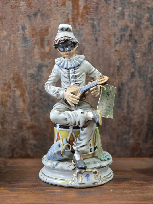 Vintage Capodimonte Porzellanfigur eines maskierten Harlekins mit Mandoline, Trommel und Goldakzenten in Frontalansicht auf Holztisch.
