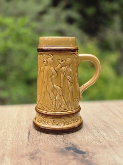 Vintage Bierkrug aus glasierter Keramik mit umlaufendem Jagdrelief, Figurenmotiv, braunen Rändern und ockerfarbener Glasur