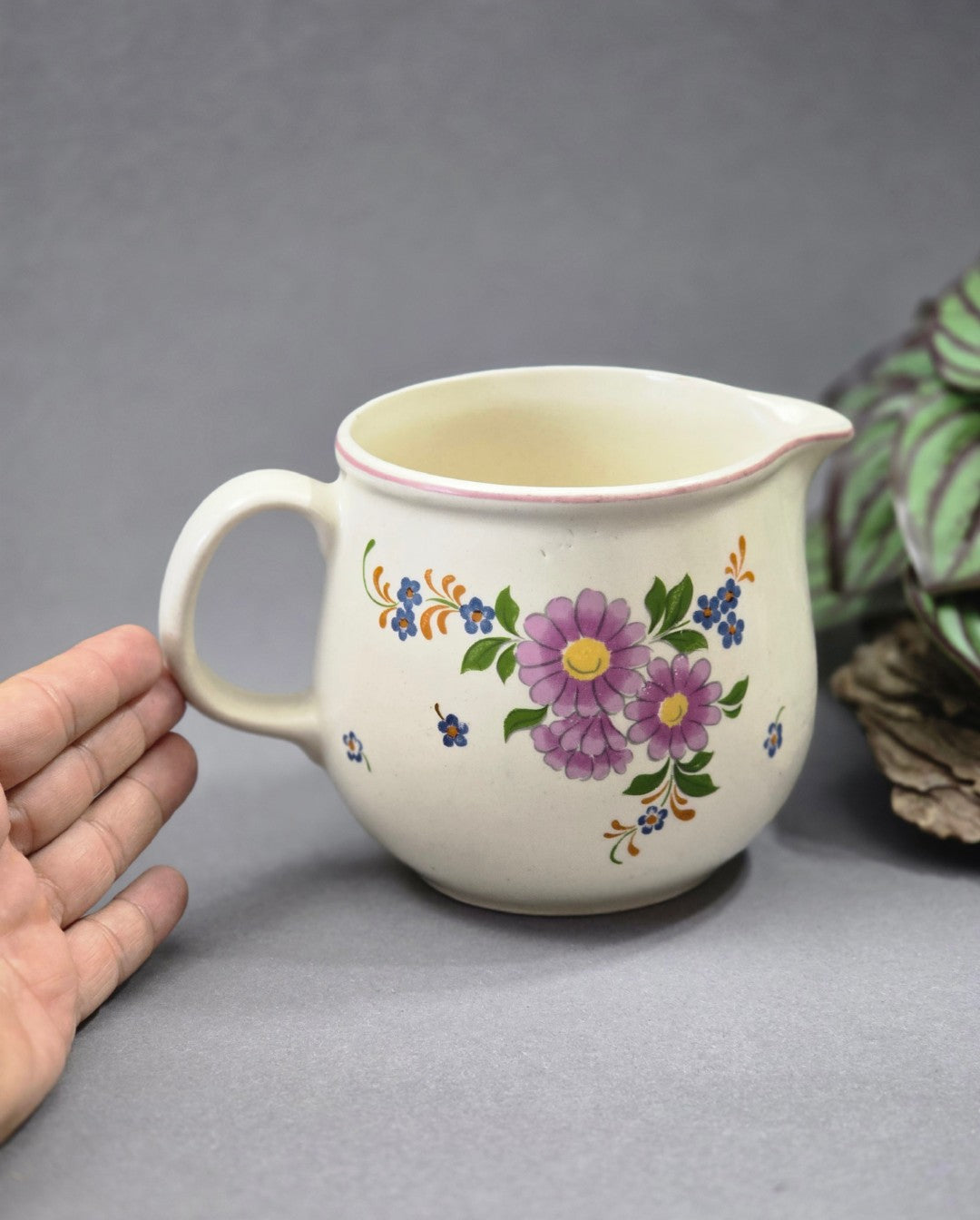 Kleines Keramikkännchen mit floralem Vintage-Dekor und Hand zum Größenvergleich, fotografiert vor neutralem grauem Hintergrund